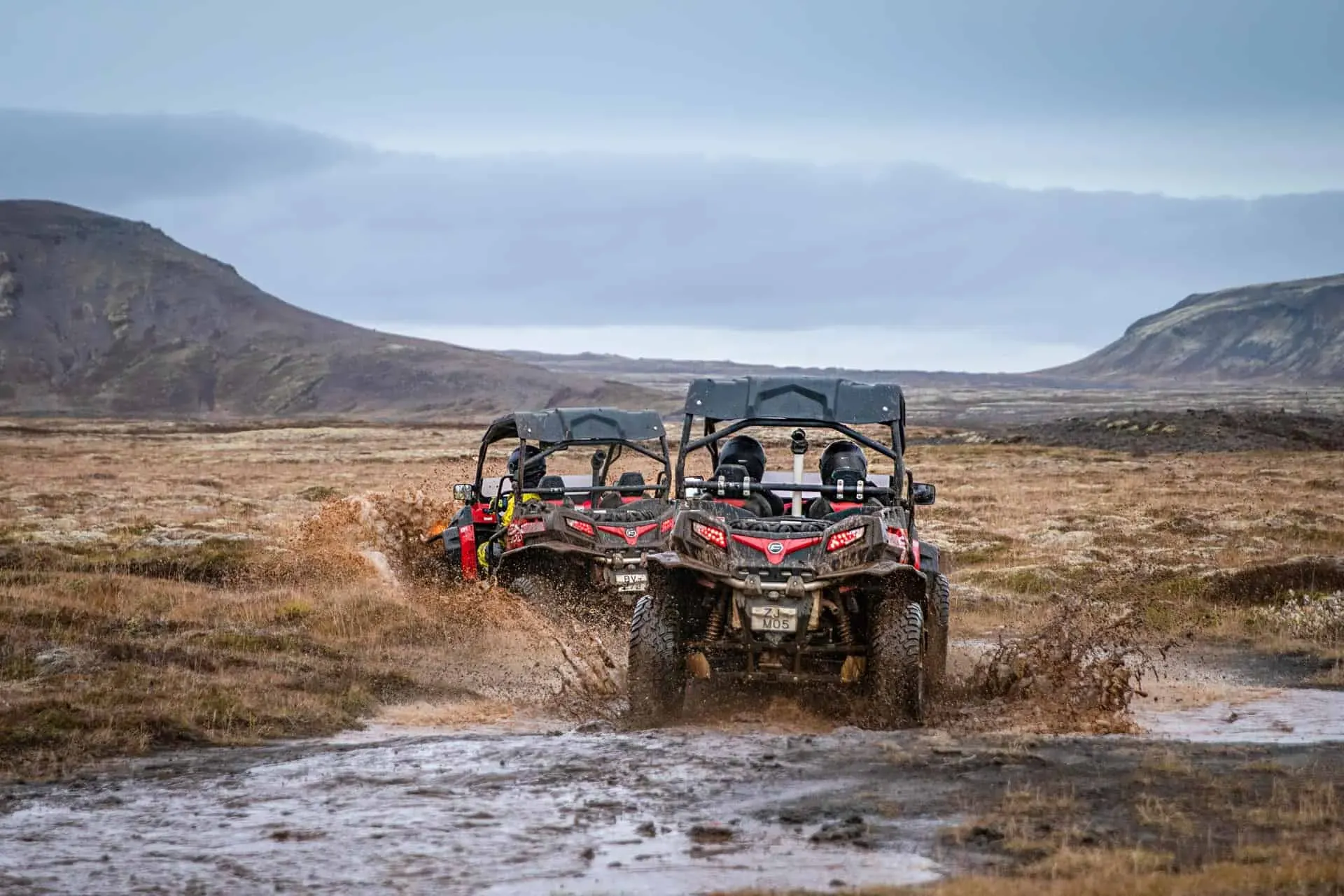 Tveir rauðir buggybílar aka í gegnum drullupoll á mosavöxnu hrauni með fjöll í baksýn